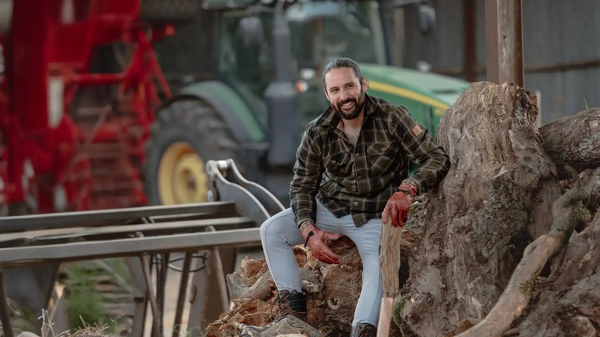 A bearded man in a plaid shirt and light-wash jeans, wearing red work gloves and holding a tool, sitting on a pile of wood.