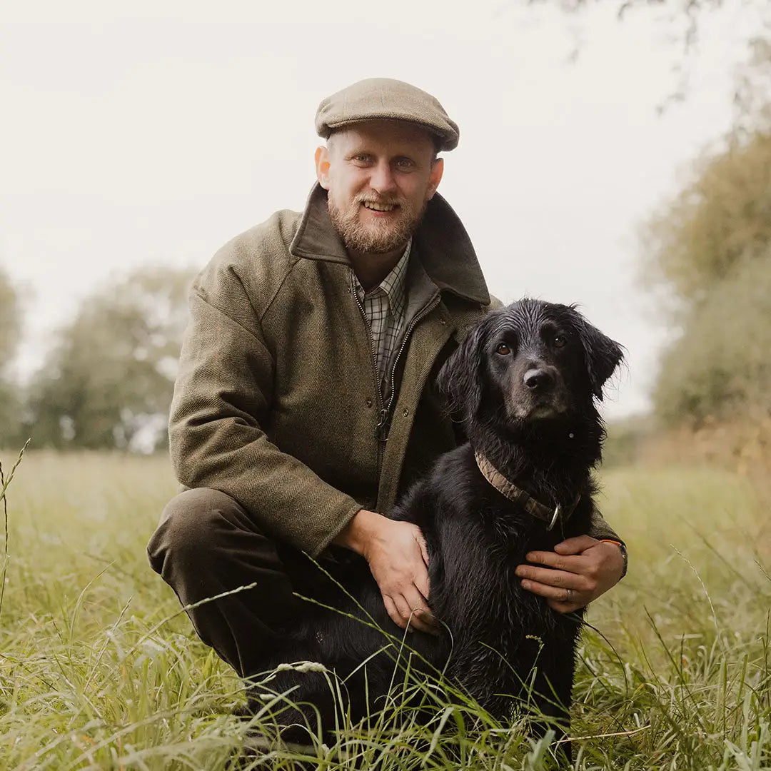 Man in a Tweed Shooting Jacket kneeling next to his black dog in a grassy field
