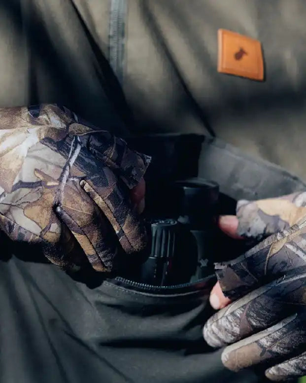 A person wearing camouflage gloves placing or retrieving a black object, possibly binoculars, from a zippered pocket of an olive green jacket.
