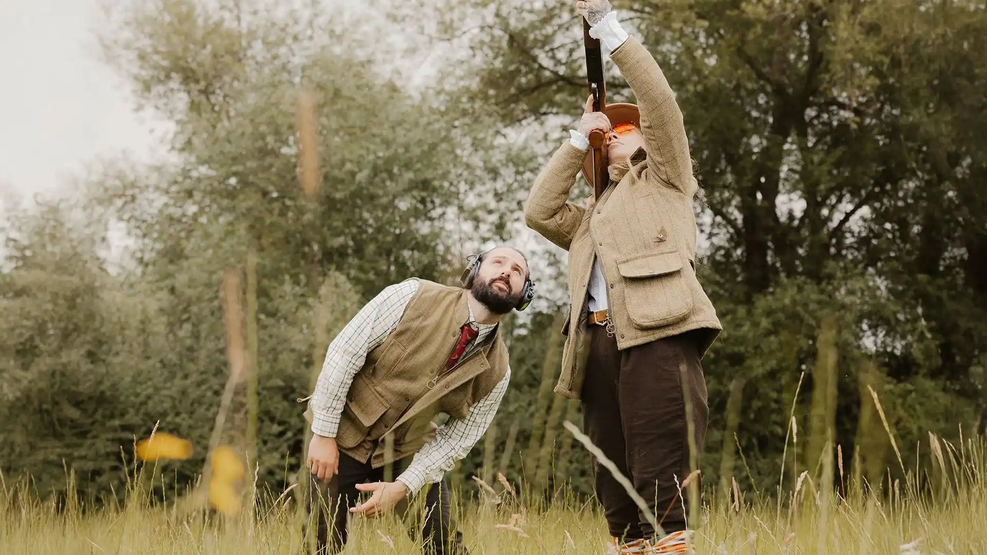 Two people dressed in shooting attire, one aiming a shotgun upwards while the other looks on.