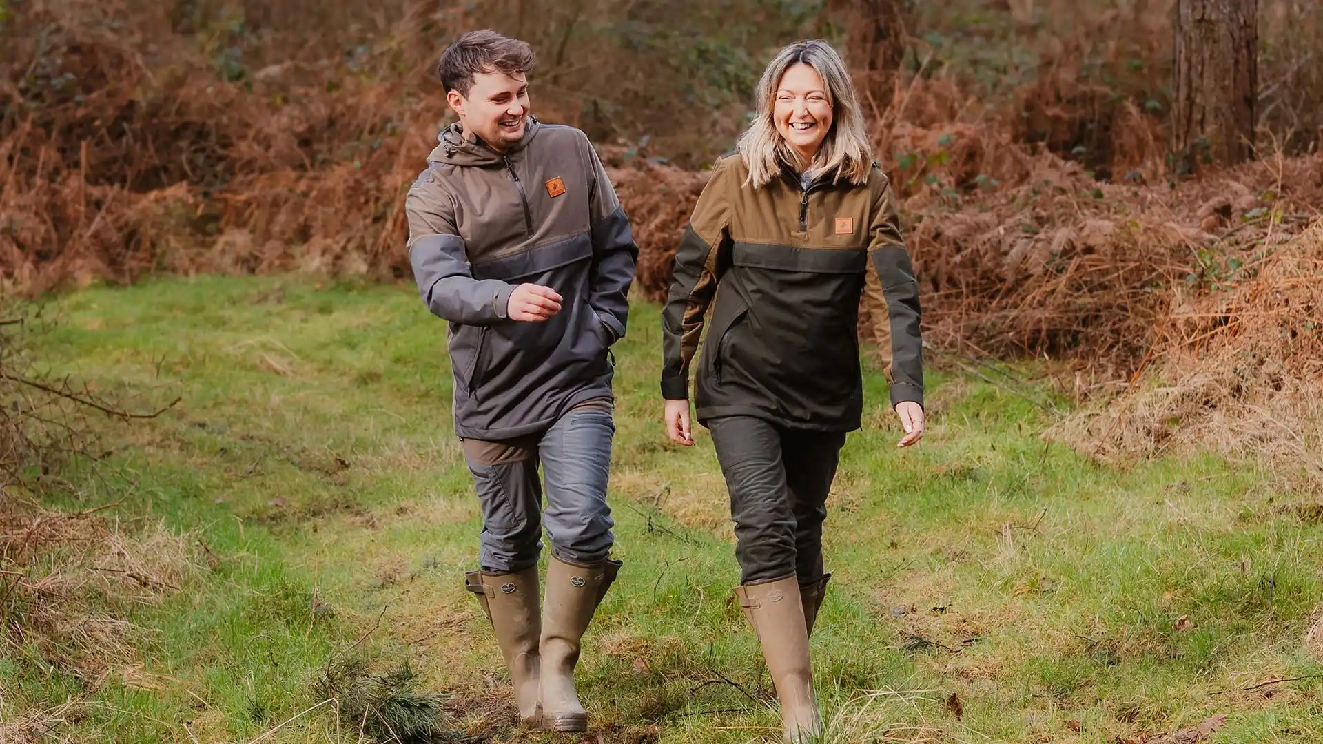 Two people wearing outdoor jackets and boots walking through a grassy area.