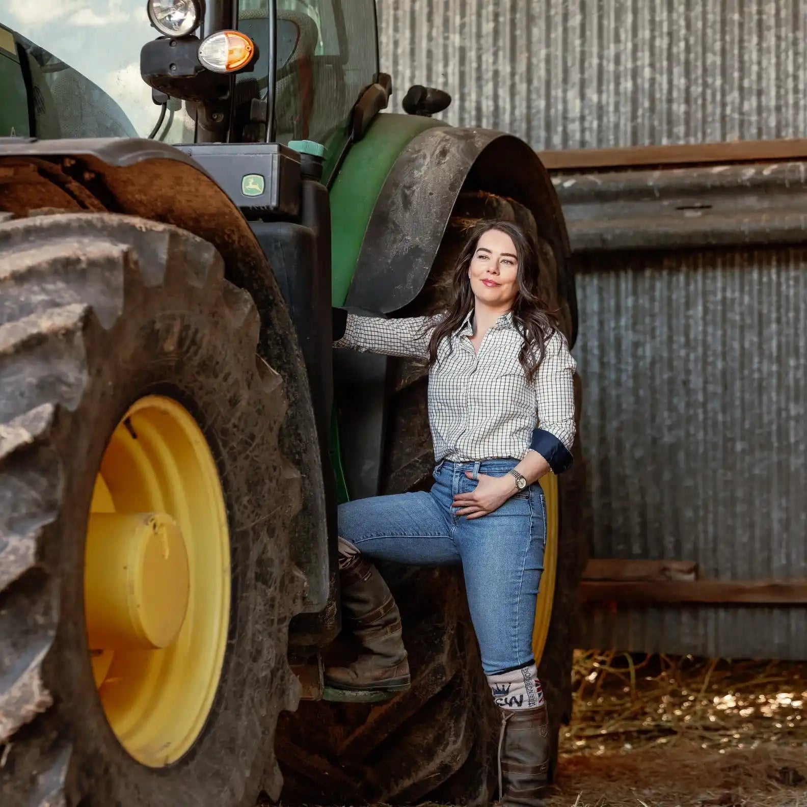 A woman in a plaid shirt, jeans, and boots posing on a large green tractor.