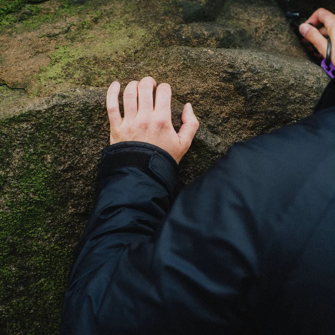 Person’s hand on mossy rock with buffalo belay jacket and purple belay device