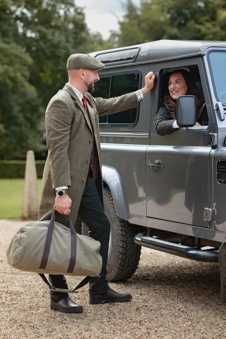 Man in tweed coat and baker boy hat talking to woman near grey SUV with khaki bag