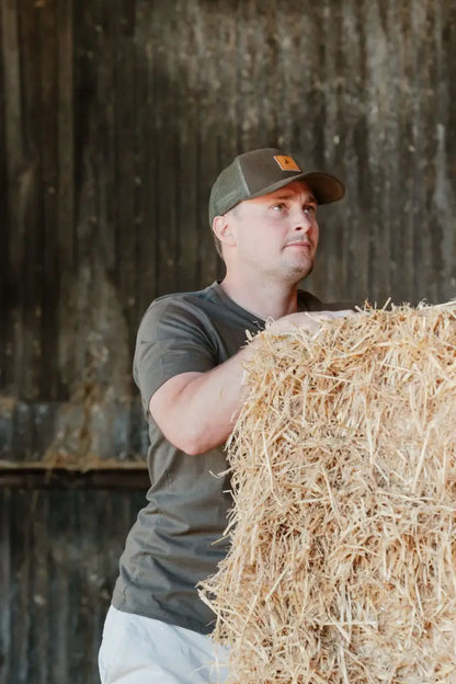 Man holding hay bale wearing New Forest Country Sport iconic trucker hat in outdoor farm scene