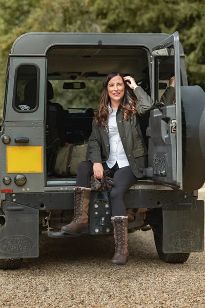 Woman in green jacket and tall brown boots sitting in grey Land Rover Defender with Forest Freya Ladies wax jacket