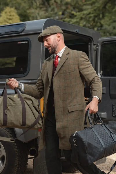 Man in forest goodwood tweed long coat carrying duffel bags outdoors