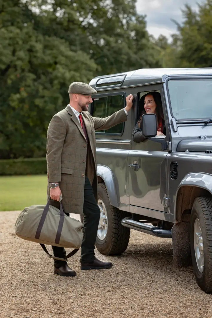 Man in forest goodwood tweed long coat talking to woman by gray SUV