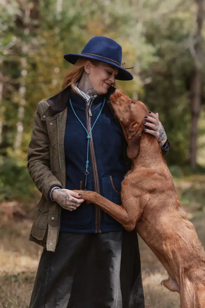 Woman in blue hat and olive jacket hugging brown dog in forest ladies fleece gilet