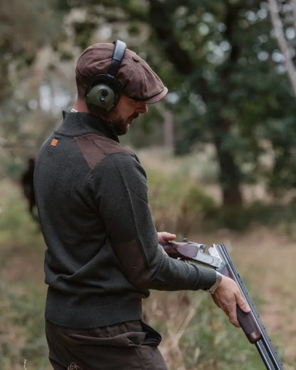 Man in forest raven zip neck jumper with shotgun, brown flat cap, and green ear defenders