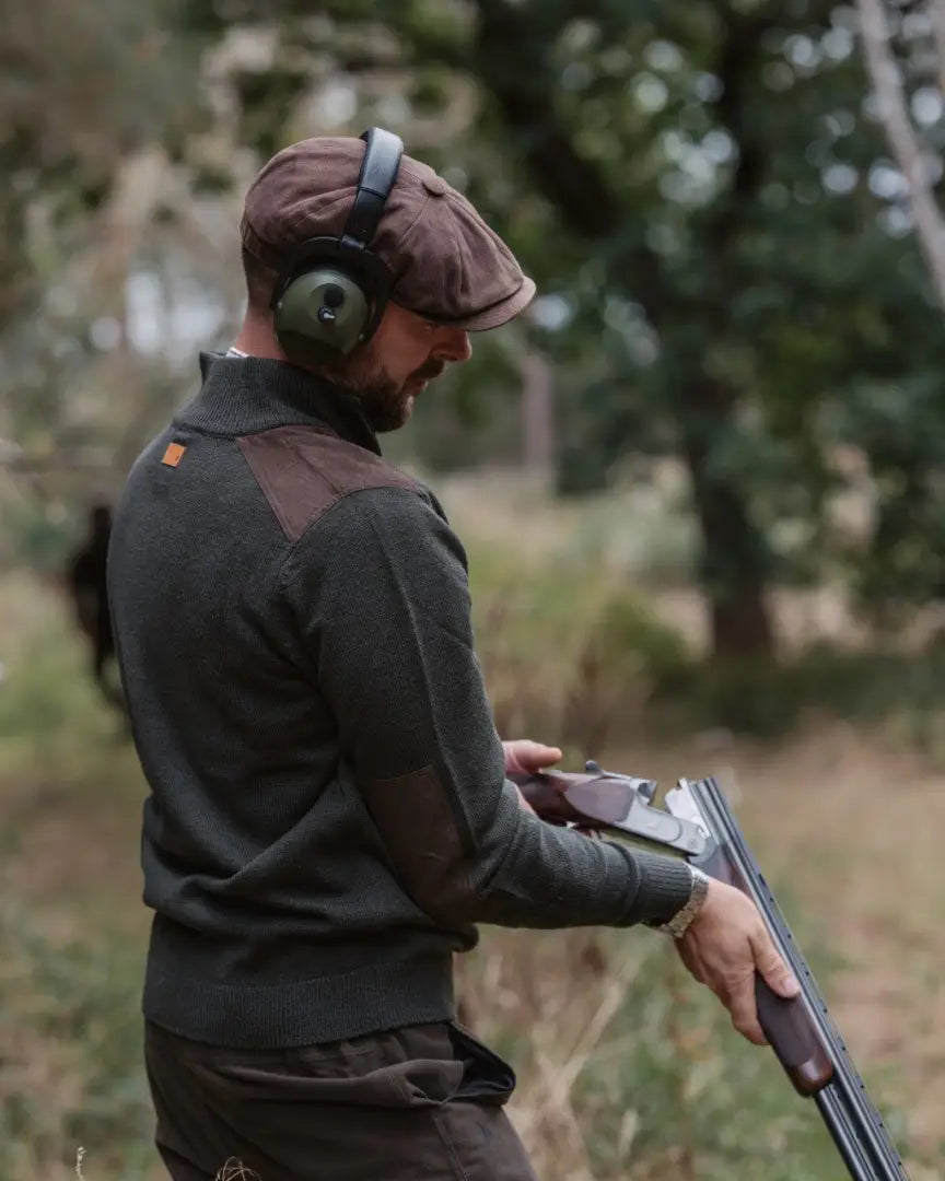 Man in forest raven zip neck jumper with shotgun, brown flat cap, and green ear defenders