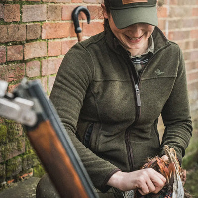 Person in a green Ivy Fleece Jacket and cap holding a shotgun outdoors