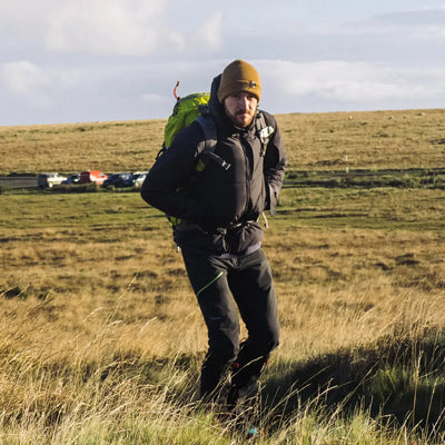 Hiker in a field wearing a Snugpak Arrowhead Jacket with a large green backpack