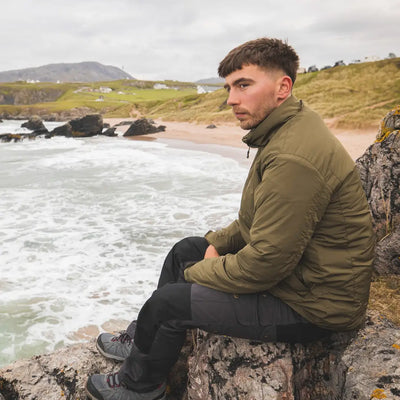 Man in a green Snugpak Arrowhead Jacket on rocky shore, part of the best selling Sleeka line