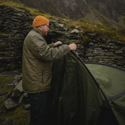 Person setting up a green tent in a rugged outdoor spot with a Snugpak Sleeka Elite Jacket